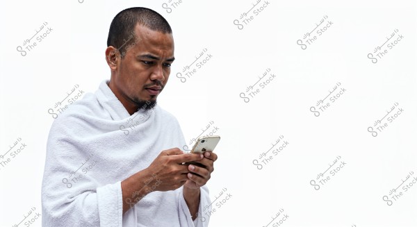 A man wearing a traditional white garment, likely in ihram attire. He is standing and using a smartphone, looking at it intently. The background is plain white, highlighting the garment and phone details.