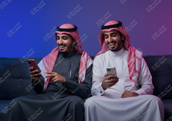 Two men sitting on a sofa, both smiling and looking at their smartphones. The man on the left is wearing a gray traditional Gulf dress and a red and white headscarf, while the man on the right is wearing a white traditional dress and a red and white headscarf. The background is lit with purple and blue colors.