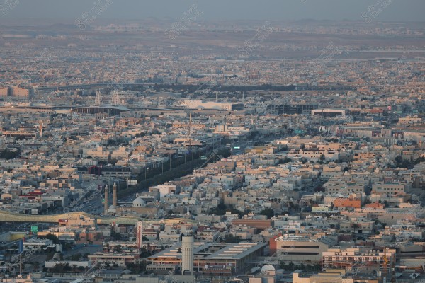 A wide view of a large city with densely packed buildings and straight streets filled with traffic, stretching across a vast area under a clear sky. In the background, there is a bridge with an intricate, modern design and hills visible on the distant horizon.