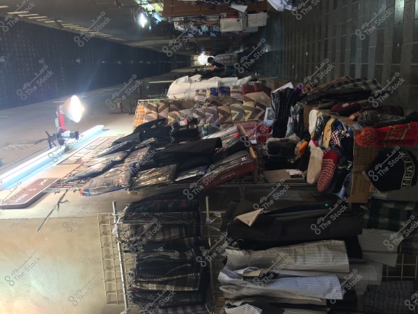 An image of a traditional market or bazaar showcasing a variety of clothing, fabrics, and pillows displayed on tables and racks. There are several lighting fixtures illuminating the area, and the floor appears to be paved with tiles. Clothing items such as jackets and caftans, as well as pillows with colorful patterns, and some knitted hats are on display.