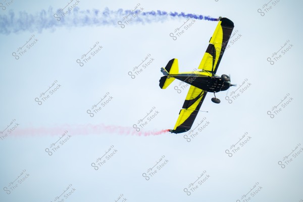 A small aircraft with striking yellow and black colors performs an aerobatic maneuver in the clear sky, leaving behind trails of blue and red smoke, adding to the visual appeal of the display.
