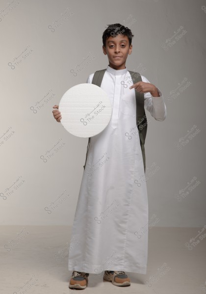 A photo of a boy wearing a traditional white thawb with a green backpack, standing against a gray background. He is holding a white circular board and pointing at himself with his finger. The clothing suggests an association with Saudi Arabia or Gulf countries.