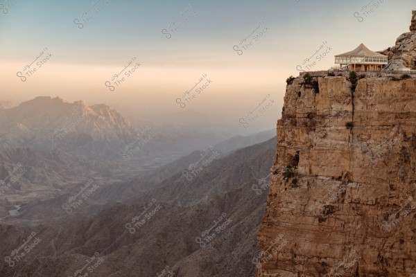 An image depicting a breathtaking view of a high cliff with a modern-designed building, possibly a restaurant, perched at its edge. It overlooks a range of mountains extending into the horizon, bathed in the golden light of sunset, enhancing the rugged beauty of the landscape.