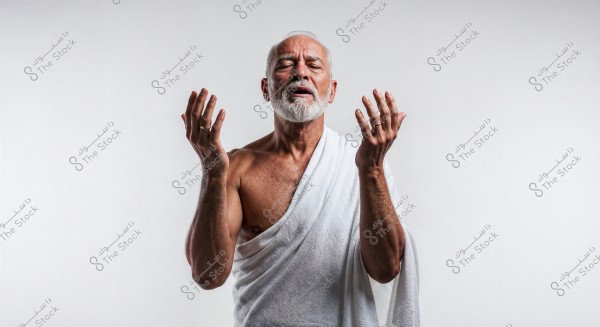 Portrait of an elderly man with a white beard, wearing a white cloth draped over one shoulder, raising his hands in a gesture of prayer or spiritual expression. The background is plain white.
