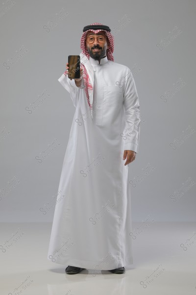 Portrait of a man wearing a white thobe and a red and white ghutra with an igal, standing and presenting a smartphone in his right hand. He appears to be from the Gulf region, possibly Saudi Arabia. The background is a neutral gray, highlighting the man\'s appearance and the phone.