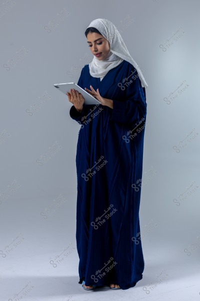 A portrait of a woman wearing a blue abaya and a white hijab, standing and using a tablet. She appears to be in a plain studio environment, looking focused as she interacts with the device.