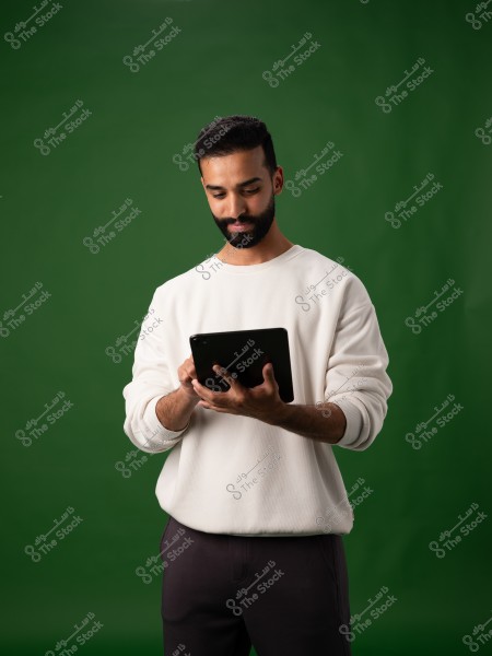 A portrait of a man holding and using a tablet, wearing a white sweater and black pants, set against a green background.
