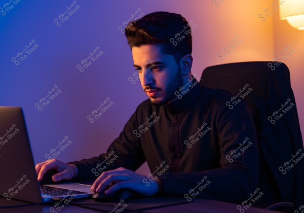 A man sitting at a desk using a laptop. He is wearing a dark-colored thobe, suggesting he may be from an Arab country such as Saudi Arabia. The room features multi-colored lighting, creating a modern and intriguing atmosphere.