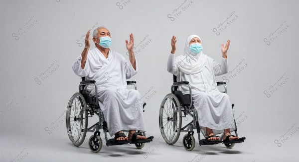 The image shows two elderly people sitting in wheelchairs, wearing white Ihram clothing and raising their hands in prayer. Both are wearing blue masks, and they are in a studio with a simple gray background.