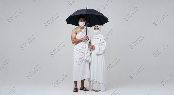 A man and a woman standing under an umbrella. The man is wearing a white ihram, and the woman is wearing a white hijab and dress. Both are wearing medical face masks. The background is plain white.