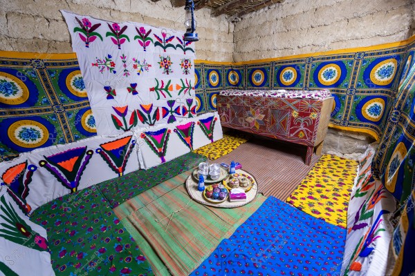 The image shows a traditional room with vibrant colors, decorated with embroidered fabrics featuring geometric patterns and flowers. Cushions on the floor are adorned with colorful embroidery. In the center of the room, there is a round tray with cups and glasses, indicating a setup for serving tea or coffee. The walls are covered with fabrics with traditional designs, and there is a distinctively designed wooden piece of furniture in the corner.