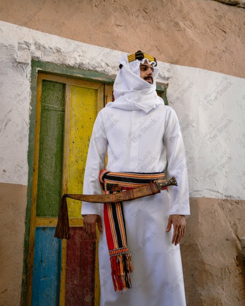 A snapshot of a Saudi man wearing a popular heritage costume on the day of the foundation stands in front of an clay building, heritage uniform, foundation day, old archaeological buildings