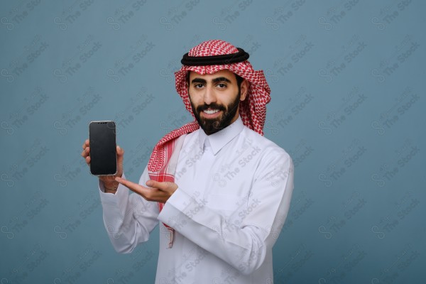 بورتريه شاب سعودي يرتدي الزي السعودي يشير إلى هاتفه ويتفاعل معه، وتظهر على ملامحه السعادة ، على خلفية سماويه