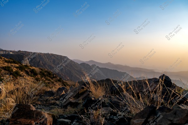 A beautiful landscape of mountains during sunrise or sunset, displaying a gradient of colors with calming blue and orange hues in the background. In the foreground, rocks and hills are covered with wild vegetation.
