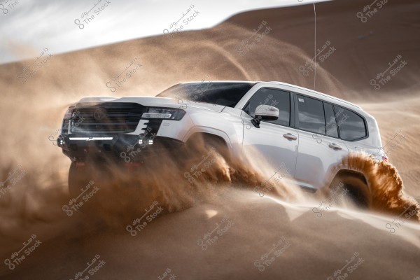 A white SUV driving swiftly across desert sand dunes, creating a dramatic effect of flying sand around the vehicle. The car is shown navigating a sandy slope, with a clear sky visible in the background.