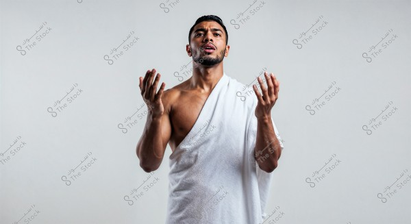 Portrait of a man standing with his hands raised in a prayer position. He is wearing a white Ihram garment that covers his left shoulder and leaves the right shoulder bare, typically worn by Muslims during the pilgrimage rituals of Hajj or Umrah. The background is plain white.