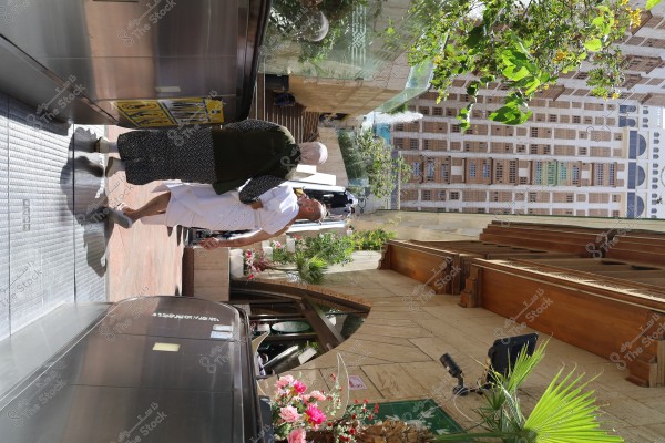 The image shows two people walking in an outdoor area next to an escalator. One person is wearing a white ihram, suggesting they might be in a location related to Mecca, Saudi Arabia. The area is surrounded by green plants and colorful flowers. In the background, there is a building with traditional design and multiple windows.