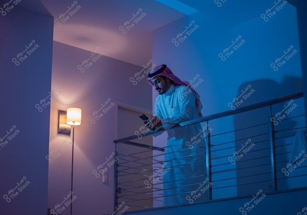 Image of a man standing on an indoor balcony lit by blue light. The man is wearing a white thobe and a red and white ghutra. He is standing beside the railing, looking at a mobile phone. In the background, there is a light-colored wall with an illuminated wall lamp.