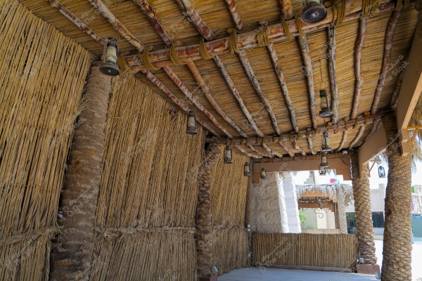 Image of a traditional structure made from palm fronds and bamboo, with palm tree trunks supporting a roof of neatly arranged wooden logs. An old lantern is hanging along the ceiling, adding a traditional touch to the place. In the background, another building made of traditional stone can be seen.