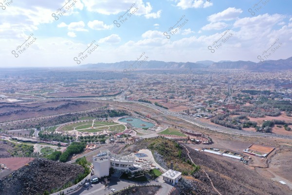 Aerial view of a sprawling city with mountainous terrain in the background and a blue sky covered with white clouds. In the foreground, there\'s a large park with a pond and several facilities. Residential buildings are spread across the vast area, surrounded by roads and other open spaces.