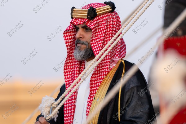 Image of a man wearing traditional Arab attire, including a red checkered headscarf (shemagh) with a black and gold agal, a white thobe, and a black bisht with gold embellishments. The photo captures the detailed expression on his face and his gray beard. The blurred background enhances the focus on the person.
