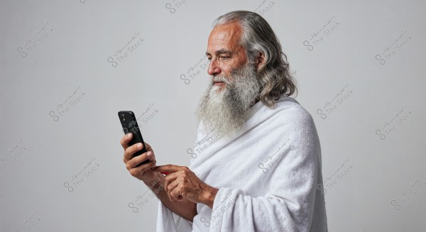 Image of an elderly man with a thick beard and long gray hair wearing a white garment resembling an ihram. The man is standing, holding a smartphone in his hand in a usage posture, with a white background, shown from the side facing left.