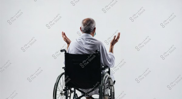 An image of an elderly man sitting in a wheelchair facing the background. The man is wearing a white robe and has his hands raised in a gesture of contemplation or prayer. The background is white, providing a calm and simple setting.
