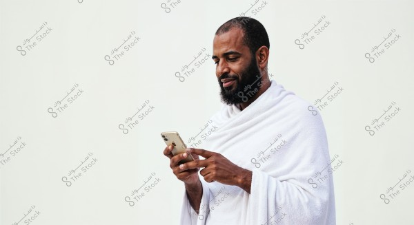 Image of a man wearing a white ihram, using a smartphone. He appears focused on the phone with a slight smile on his face. The background is plain white and simple.