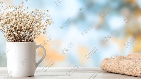 A large white mug holds a bunch of small white dried flowers, placed on a wooden surface next to a piece of brown fabric. The background is blurred with soft blue and yellow hues.