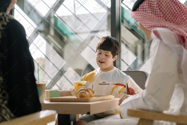 طفل "من ذوي الاعاقة" يجلس على طاولة مقهى مع اثنين من البالغين، يتناول عصير ومعجنات.