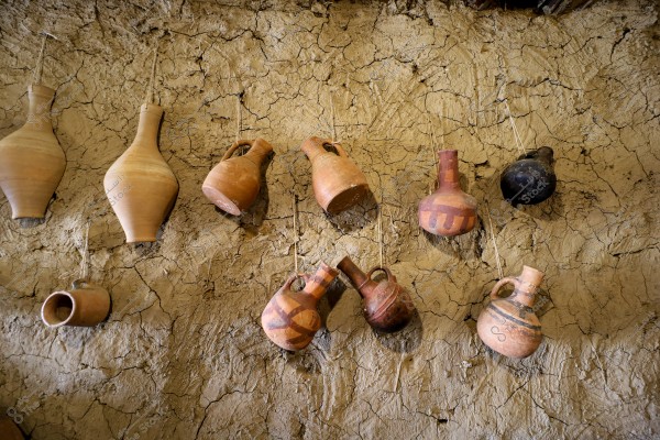 The image shows a collection of ancient clay pots hanging on a mud wall with a natural cracked pattern. The pots vary in size and shape, featuring earthy and brown tones with some simple decorations. The background wall adds a traditional touch with its earthy texture and distinctive cracks.