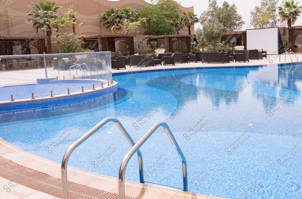 An image of an outdoor swimming pool at a hotel or resort, enclosed by a glass fence. The photo shows the clear blue water of the pool with shiny curved metal steps leading into it. In the background, there is a luxurious seating area with black chairs and palm trees, along with other greenery and a large display screen.