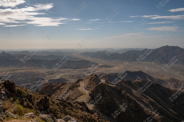 A landscape view of mountains and plains stretching into a distant horizon under a blue sky with some light clouds. A winding road crosses the mountainous area with some patches of vegetation on the rocks in the foreground.