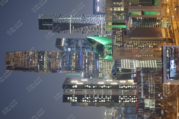 An image showing a city skyline in the evening with tall, modern buildings glowing with lights. Some buildings feature geometric designs and green and blue lights, giving a contemporary flair to the night scene. The dark sky enhances the visibility of the lights on the buildings.