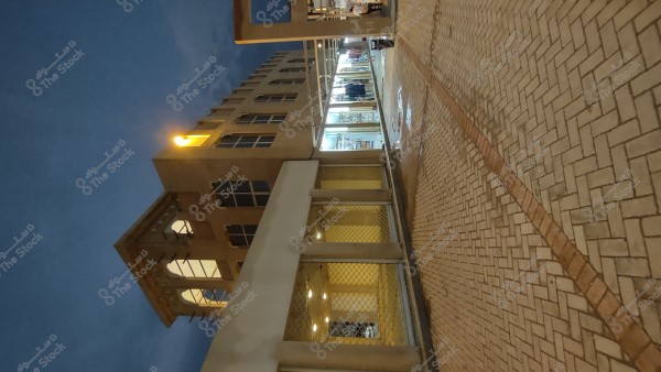 The image shows a section of a multi-story building with glass windows and exterior lights. There are also lit shops on the right side of the image, and a paved sidewalk running along the building. The sky is dark, suggesting it is evening.