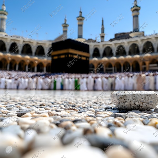 Image of a ground surface covered with pebbles, with the Kaaba in the background at Mecca. Crowds of people are gathered around the Kaaba, wearing traditional white clothing. The minarets of the Grand Mosque are visible against a clear blue sky.