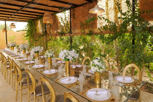Image of a long dining table illuminated by natural light, set in an outdoor space surrounded by green plants. The table has a glass top and is accompanied by gold chairs. It is adorned with white plates, folded napkins, and golden cutlery, while golden vases with white flowers decorate the table. The ceiling is made of wood and bamboo, with several hanging lamps.