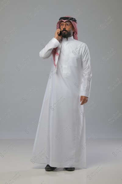 A man wearing traditional Saudi attire, consisting of a white thobe and a red checkered ghutra with a black agal. The man is standing in a natural pose while using a mobile phone. The background is gray and neutral.