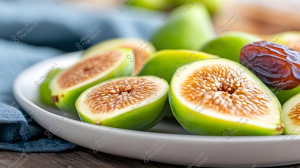 صورة لصحن يحتوي على تين طازج مقطّع إلى نصفين يظهر لونه الأخضر الخارجي ولحم التين الداخلي اللبني مع البذور. يوجد أيضًا تمر بني لامع بجانب التين في الطبق. خلفية الصورة تحتوي على قماش أزرق وطاولة خشبية.