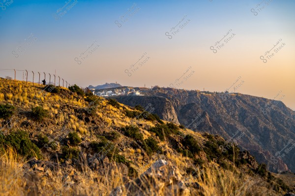 A natural landscape of a mountain covered with scattered grass and greenery during sunset. In the background, white buildings are visible along the horizon on the mountain. The sky is a mix of blue and orange, suggesting a beautiful sunset.