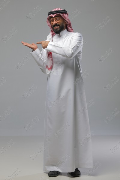 Image of a man standing and smiling, pointing with one hand. He is wearing traditional Saudi attire, which consists of a white thobe and a red and white checkered headscarf (ghutra) with a black band (agal). The background is a neutral light gray.