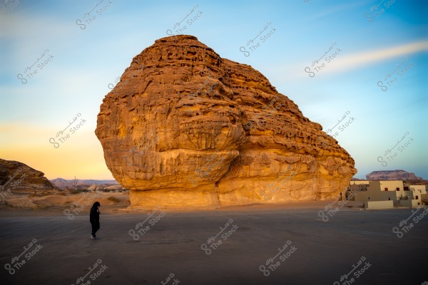 The image shows a large rock formation with prominent rocky textures, displaying vibrant colors under a blue sky. A person is walking near the rock, dressed in black clothing, possibly indicating local attire. Some simple buildings appear in the background, suggesting a small residential area.