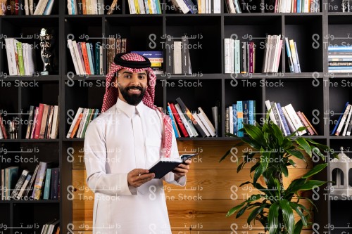 رجل سعودي يرتدي الزي السعودي التقليدي يقف في المكتبة مبتسمًا بينما يتصفح أحد الكتب