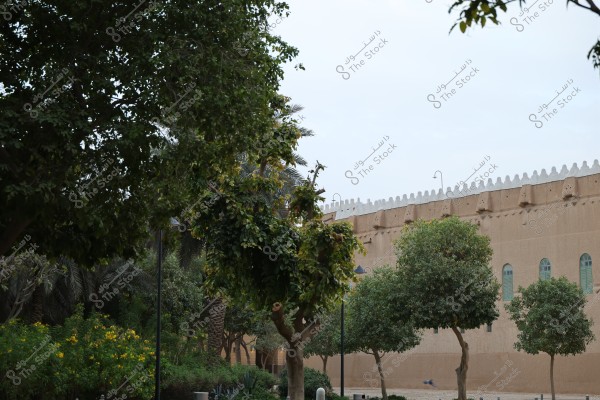The image shows a garden with dense green trees and yellow flowers. In the background, there is a wall of a traditional mud building with arched windows of green glass and decorations at the top. Trees and plants line the path, adding natural beauty to the scene.