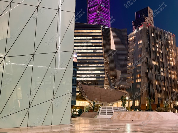 A photo of a modern building complex at night, with the buildings\' lights illuminated. The structure in the foreground has an angular design with intersecting lines. In the background, a tower with purple lighting is visible, while other buildings feature prominently lit windows. There is also a curved architectural element in the center with scattered greenery.