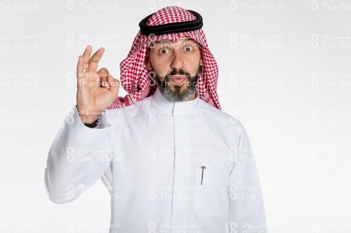 بورتريه رجل سعودي يقف على خلفية بيضاء، يرتدي الثوب، يقف امام الكاميرا مندهش ويقوم بعمل حركات بيديه، تعبير على اتمام شيء او الاتفاق مع شيء