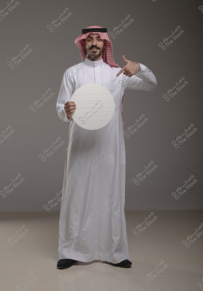 Portrait of a man standing in a studio wearing traditional Saudi attire, including a thobe, ghutra, and agal. He is holding a white circle and appears to be pointing at it with his other hand. The background is light gray.