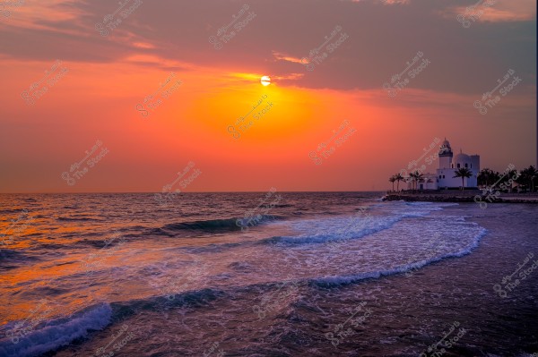 غروب الشمس فوق البحر الأحمر مع مبنى ومجموعة من الأشجار في الخلفية.