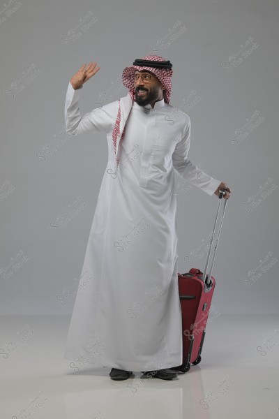 Image of a man wearing a white thobe and traditional headscarf, possibly from Saudi Arabia. He is standing next to a red suitcase, smiling and waving.