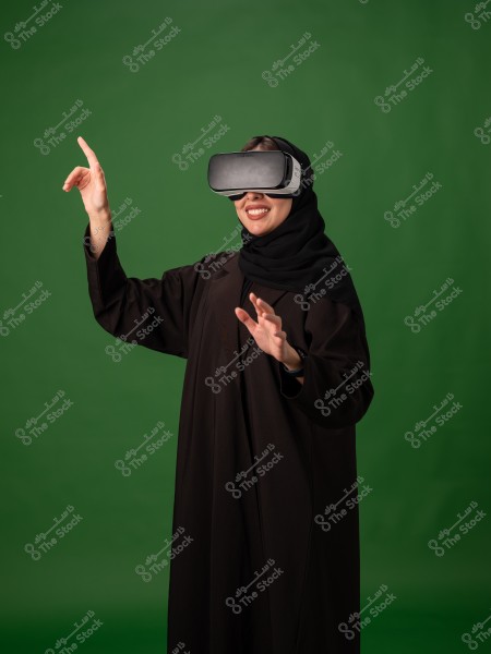 A woman wearing an abaya and virtual reality glasses, standing in front of a green background.